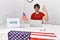 Young hispanic man at political election sitting by ballot smiling positive doing ok sign with hand and fingers