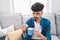 Young hispanic man pouring vitamin on coffee sitting on sofa at home