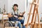 Young hispanic man sitting on wheelchair painting at art studio looking away to side with smile on face, natural expression