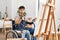 Young hispanic man sitting on wheelchair painting at art studio looking at the camera blowing a kiss with hand on air being lovely