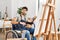 Young hispanic man sitting on wheelchair painting at art studio smiling looking to the side and staring away thinking