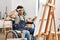 Young hispanic man sitting on wheelchair painting at art studio very happy and smiling looking far away with hand over head