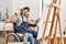 Young hispanic man sitting on wheelchair painting at art studio waiving saying hello happy and smiling, friendly welcome gesture