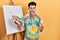 Young hispanic man standing drawing with palette by painter easel stand angry and mad raising fist frustrated and furious while