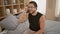 Young hispanic man taking glasses off sitting on bed smiling at bedroom