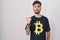 Young hispanic man with tattoos wearing bitcoin t shirt surprised pointing with hand finger to the side, open mouth amazed