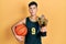 Young hispanic man wearing basketball uniform holding ball and prize making fish face with mouth and squinting eyes, crazy and