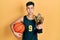 Young hispanic man wearing basketball uniform holding ball and prize puffing cheeks with funny face