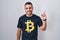 Young hispanic man wearing bitcoin t shirt with a big smile on face, pointing with hand finger to the side looking at the camera