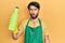 Young hispanic man wearing cleaner apron holding cleaning product scared and amazed with open mouth for surprise, disbelief face