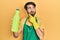 Young hispanic man wearing cleaner apron holding cleaning product serious face thinking about question with hand on chin,