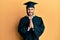 Young hispanic man wearing graduation cap and ceremony robe praying with hands together asking for forgiveness smiling confident