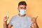 Young hispanic man wearing medical mask holding syringe pointing thumb up to the side smiling happy with open mouth