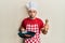 Young hispanic man wearing professional cook uniform and hat cooking beef making fish face with mouth and squinting eyes, crazy