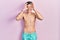 Young hispanic man wearing swimwear shirtless doing ok gesture like binoculars sticking tongue out, eyes looking through fingers