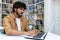 Young hispanic student studying inside university campus academic library among bookshelves, man smiling and using