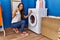 Young hispanic woman doing laundry choosing washing machine program smiling happy and positive, thumb up doing excellent and