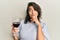 Young hispanic woman drinking a glass of red wine serious face thinking about question with hand on chin, thoughtful about