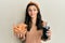 Young hispanic woman eating chicken wings and drinking soda smiling looking to the side and staring away thinking