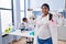 Young hispanic woman expecting a baby working at scientist laboratory looking confident with smile on face, pointing oneself with