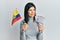 Young hispanic woman holding colombia flag and colombian pesos banknotes smiling looking to the side and staring away thinking