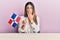 Young hispanic woman holding dominican republic flag sitting on the table covering mouth with hand, shocked and afraid for mistake