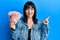 Young hispanic woman holding icelandic krona banknotes smiling happy pointing with hand and finger to the side