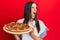 Young hispanic woman holding italian pizza angry and mad screaming frustrated and furious, shouting with anger