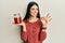 Young hispanic woman holding jar with honey doing ok sign with fingers, smiling friendly gesturing excellent symbol