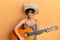 Young hispanic woman holding mexican hat playing classical guitar clueless and confused expression