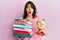 Young hispanic woman holding a pile of books and world ball afraid and shocked with surprise and amazed expression, fear and