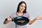 Young hispanic woman holding skillet with beef steak celebrating achievement with happy smile and winner expression with raised