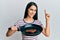 Young hispanic woman holding skillet with beef steak smiling with an idea or question pointing finger with happy face, number one