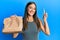 Young hispanic woman holding take away paper bag surprised with an idea or question pointing finger with happy face, number one