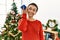 Young hispanic woman listening smartphone audio standing by christmas tree at home