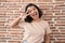 Young hispanic woman standing over bricks wall doing peace symbol with fingers over face, smiling cheerful showing victory