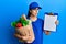 Young hispanic woman wearing courier uniform with groceries from supermarket and clipboard clueless and confused expression