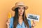 Young hispanic woman wearing hat holding ice cream and hello summer paper clueless and confused expression