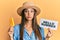 Young hispanic woman wearing hat holding ice cream and hello summer paper puffing cheeks with funny face