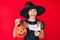 Young hispanic woman wearing witch costume holding pumpkin and happy halloween message angry and mad screaming frustrated and