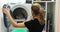 Young housewife unloading washing machine and putting clean clothes in laundry basket. Woman picking up clothes out of