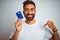 Young indian customer man holding credit card standing over isolated white background with surprise face pointing finger to