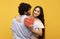 Young indian lovers hugging on yellow background, lady holding heart shaped gift box and smiling at camera