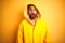 Young indian man wearing raincoat with hood standing over isolated yellow background afraid and shocked with surprise expression,