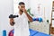 Young indian physiotherapist holding therapy massage gun at wellness center smelling something stinky and disgusting, intolerable