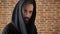 Young interesting man with beard in hood is watching at camera, goes away, brick background