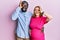 Young interracial couple expecting a baby, touching pregnant belly doing ok gesture with hand smiling, eye looking through fingers