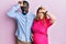 Young interracial couple expecting a baby, touching pregnant belly making fun of people with fingers on forehead doing loser