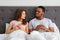 Young interracial couple with smartphones sitting in bed, stuck in gadgets, looking at each other with suspicion