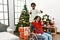 Young interracial couple with woman sitting on wheelchair by christmas tree approving doing positive gesture with hand, thumbs up